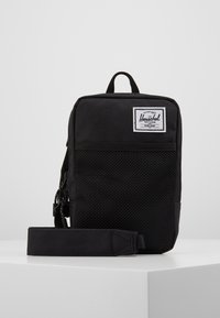 Sac bandoulière compact Herschel noir avec une poche avant en maille, une sangle réglable, une poignée supérieure et une étiquette de logo de marque.