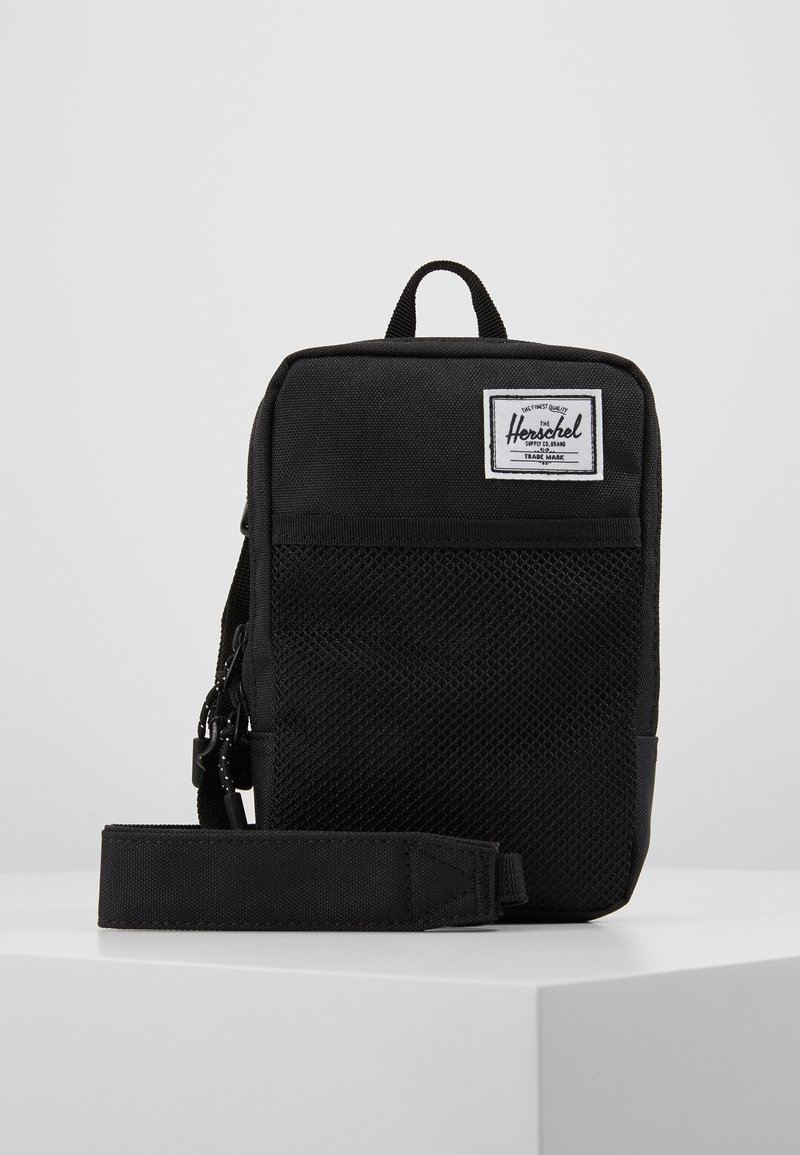 Sac bandoulière compact Herschel noir avec une poche avant en maille, une sangle réglable, une poignée supérieure et une étiquette de logo de marque.