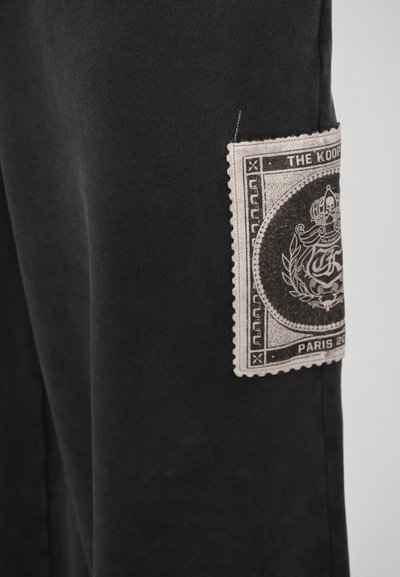 Sorte bukser lavet af blødt stof med et fremtrædende tekstureret klistermærke med et vintage design og dekorativ kant på siden.