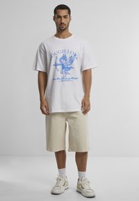 White cotton t-shirt featuring blue graphic print of a winged figure with "TOUGH LOVE" text. Paired with beige knee-length shorts and light sneakers.