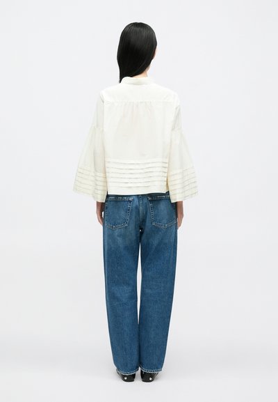 Kvinde med langt sort hår iført en hvid bluse med plisserede vide ærmer og blå jeans med lige ben, set bagfra.