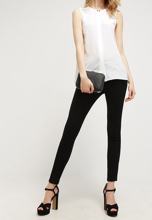 Blusa bianca senza maniche con apertura a bottoni, abbinata a pantaloni neri slim-fit e scarpe con tacco alto e cinturino nero. Piccola pochette nera.