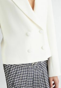Witte dubbelzijdige blazer van textuurstof, met grote ronde knopen. Draag het over een geplooide marineblauwe en beige gingham rok.