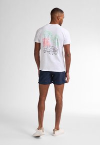T-shirt bianco di cotone con una grafica colorata di palme e un camion. Indossato con pantaloni corti blu navy e scarpe di colore chiaro.