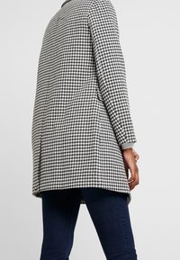 Cappotto a motivo houndstooth in bianco e nero, con vestibilità ampia, maniche lunghe e dettagli a coste grigi ai polsini.