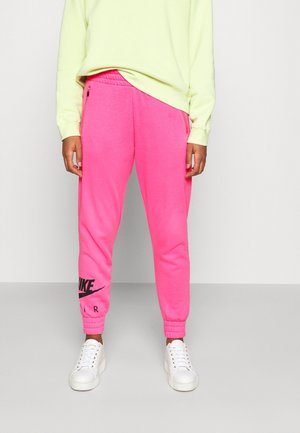 Persoon die felroze Nike Air sweatpants met ritssluitzakken, een lichtgele trui en witte sneakers draagt en op een grijze vloer staat.