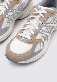 Zapatilla de malla blanca con acentos de ante gris y beige, con suela gruesa, cordones planos y detalles reflectantes en el costado.