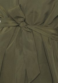 Látka olivově zelené barvy s hladkou texturou, zdobená zavázanou mašlí v pase a viditelnými stehům.