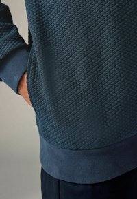 Sudadera de un azul oscuro con textura, confeccionada con una tela de patrón de diamante, con puños y dobladillo acanalados. Presenta un bolsillo lateral con ribete de tejido suave.