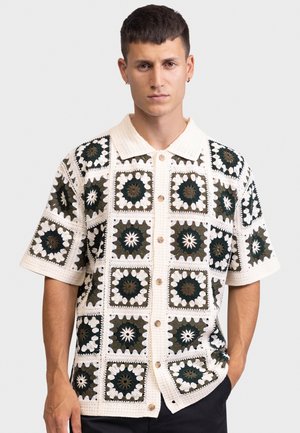 Bărbat cu păr scurt și închis la culoare, purtând o cămașă albă cu mâneci scurte din croșetat, cu modele pătrate florale negre și verzi, stând cu o mână în buzunar.