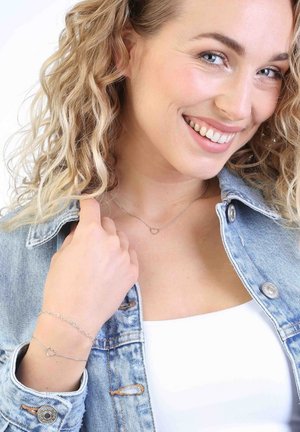 Silberne Herz-Halskette und Armband an einer Person, die eine Jeansjacke über einem weißen Oberteil trägt; lockige Haare; Akzentdetails im Schmuck.
