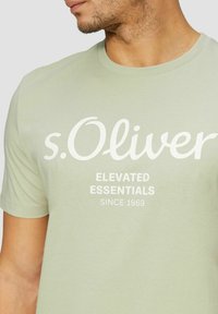 Light green cotton t-shirt with short sleeves, featuring white printed text: "s.Oliver," "ELEVATED ESSENTIALS," and "SINCE 1969."