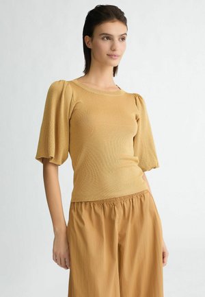 Maglione color senape a maglia con maniche a sbuffo, caratterizzato da un collo rotondo e una silhouette aderente. Abbinato a pantaloni beige morbidi.