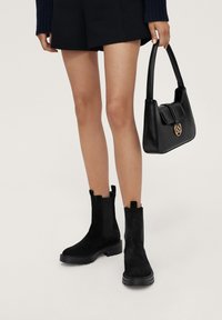 Bottes de cheville en suede noir avec une finition texturée, des panneaux latéraux élastiques et des languettes de tirage. Un sac à main noir avec un emblème doré et une fermeture à rabat les complète.