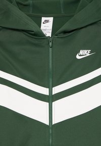 Zaļš Nike kapuce ar aizdari, kas ir balta, piedāvā divas baltas ševron formas svītras uz krūtīm un logo kreisajā pusē.