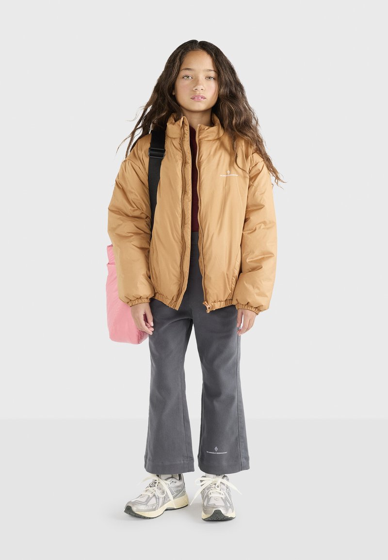 Giacca a vento marrone chiaro, pantaloni grigi a gamba larga e sneakers grigi. Zaino rosa indossato a spalla. Capelli lunghi e mossi. Sfondo semplice.
