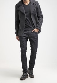 Hombre con abrigo corto gris oscuro sobre suéter negro de cuello en V, jeans oscuros de corte ajustado y zapatos de vestir de cuero negros, de pie con una mano en el bolsillo.