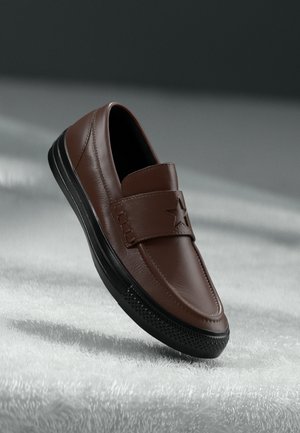 Brūns ādas slip-on apavi ar mīkstinātu siksnu, kas rotāta ar zvaigznes detaļu, melna gumijas zole un strukturēti akcenti sānos.