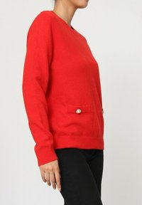 Pull rouge à manches longues, col rond et deux poches ornées de boutons. Fabriqué en tissu doux et texturé, design ajusté.
