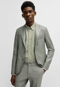 Abito grigio su misura con una sottile texture, dotato di due bottoni, abbinato a una camicia verde chiaro con bottoni. Entrambi hanno una vestibilità moderna.