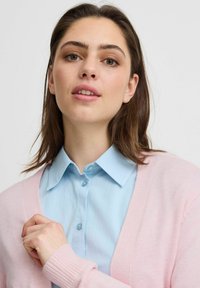 Cardigan rosa chiaro sopra una camicia colletto azzurro. Il cardigan presenta una texture morbida e un design con apertura frontale e polsini a coste.