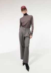 Vrouw in een grijze coltrui, wijde grijze broek, zwarte puntige schoenen en een bordeauxrode pet, staand tegen een witte achtergrond.