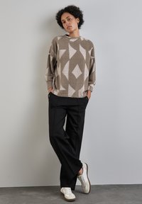 Světle hnědý svetr s diamantovým vzorem, černé široké kalhoty a bílé tenisky. Měkká textilie, volný střih, podrobné švy a kapsy.