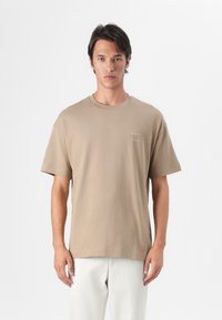 Beige bomulds t-shirt med rund halsudskæring og korte ærmer. Har et lille logo på venstre side af brystet. Glat tekstur, afslappet pasform.