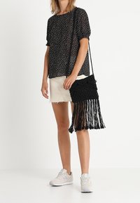 Černá makramé crossbody taška s třásněmi, zdobená složitými uzlovými vzory. Modelka má na sobě top s puntíky a bílé kraťasy, doplněné teniskami.