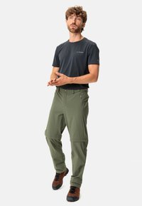 Männliches Outdoor-Ensemble mit einem taillierten schwarzen Kurzarmhemd und wandelbaren grünen Hosen mit Reißverschlusstaschen und robuster Stoffqualität.