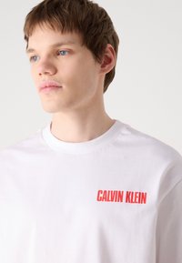 Hvit bomull t-skjorte med rund hals og korte ermer, med rødt "CALVIN KLEIN" tekstlogo på venstre brystområde.