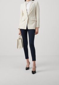 Blazer blanco a medida, camisa blanca, pantalones ajustados azul marino, zapatos de charol negros, sosteniendo un bolso texturizado color crema. Líneas limpias y diseño estructurado.