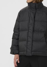 Sort puffer-jakke med høj krave, med quiltede paneler, en lige silhuet og skjult frontlukning. Glat, skinnende tekstur.