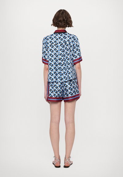 Cămașă cu mâneci scurte de culoare albastră și pantaloni scurți asortați cu un model geometric, margine roșie și o croială relaxată. Materialul pare ușor și neted.