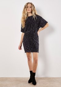 Schwarzes Minikleid mit lockerer Passform, das eine glitzernde Textur und kurze Ärmel aufweist. Stilisiert mit schwarzen Knöchelstiefeln für einen Kontrast.