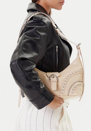 Mujer con chaqueta de cuero negra y pantalones blancos sostiene un bolso beige de hombro con detalles de tachuelas plateadas y hebilla.