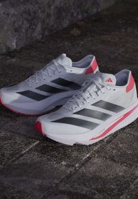 Witte sport schoenen met grijze drie strepen, rode accenten op de hiel en de tussenzool, lichtgewicht mesh bovenwerk en een gestructureerde zool voor grip.