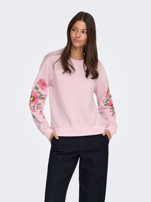 Woman with long brown hair wearing a light pink sweatshirt with pink floral embroidery on sleeves and dark pants, standing with hands in pockets.