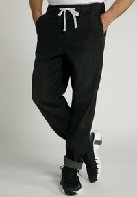 Zwarte joggingsbroek met elastische tailleband en witte trekkoord, voorzien van zijzakken en een ontspannen fit, gecombineerd met zwarte sneakers.