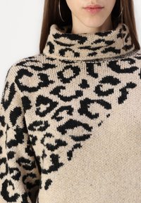 Pull en tricot beige avec un motif animal noir, doté d'un col haut et d'une texture structurée. Accent sur les couleurs et motifs contrastants.
