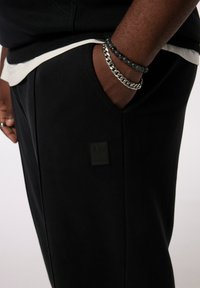 Sorte sweatpants med en glat tekstur, der har en sidelomme og et rektangulært mærke på venstre lår. Casual design med et hvidt accent.