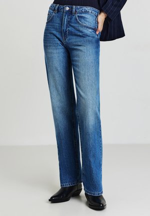 Jeans in denim blu con un design a gamba dritta, texture sbiadita, cinque tasche e chiusura con bottone. Indossati con stivaletti neri alla caviglia.