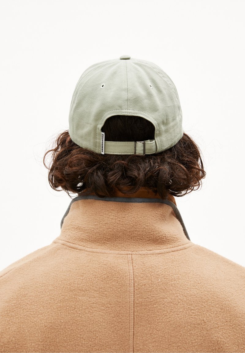 Personne aux cheveux bouclés portant une veste en polaire beige et une casquette de baseball vert clair, vue de dos sur un fond uni.