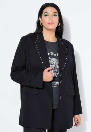 Kvinde med langt mørkt hår iført en sort frakke med nitter, grafisk T-shirt, guldringeøreringe og grøn øjenskygge, stående mod en hvid baggrund.