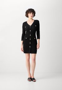 Robe en tricot noire avec un décolleté en V, des manches trois-quarts et une fermeture par boutons. Comprend deux poches frontales, chacune ornée d'un bouton décoratif.