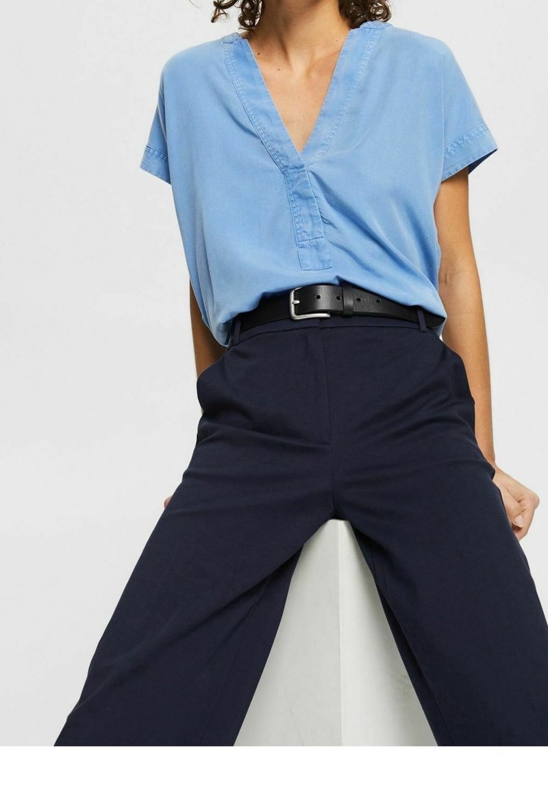 Personne portant un t-shirt bleu clair à manches courtes et col en V, rentré dans un pantalon taille haute bleu marine foncé avec une ceinture noire.