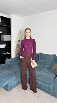 Kehas sobiv marooni värvi ribaline sviiter, kõrge talje ja laiad pruunid püksid ning beež risti üle õla kott; seisab tekstuurilisel türkiissinisel diivanil.