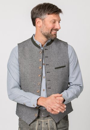 Mann in traditioneller bayerischer Tracht mit grauer Weste, gestreiftem Hemd und bestickter Lederhose, lächelnd und nach rechts schauend.