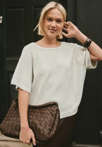 Blonde woman smiling, wearing a sparkly cream short-sleeve sweater, holding a large brown woven clutch, and touching her hair with one hand.