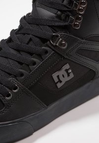 Svart høyhals sneaker med lær- og tekstiloverdel, teksturert overflate, metalløyer og en fremtredende logo på siden.